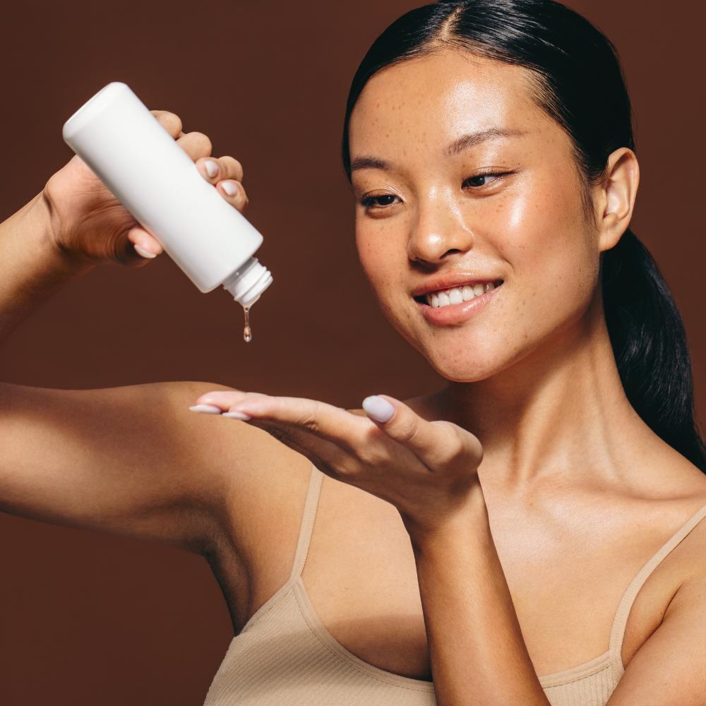 Woman applying a white bottle of skincare product to her arm against a brown background

