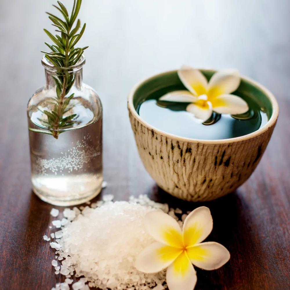 Small bottle with water and plant, ceramic bowl with flower, and salt on a wooden surface.


