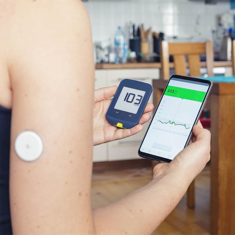 Person monitoring blood sugar levels using a device and smartphone app in a kitchen setting.

